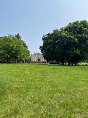 A large, historic building with a symmetrical facade, surrounded by a lush green field with scattered wildflowers and trees.