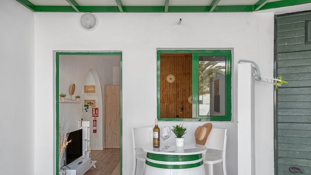 A room with a TV, a small table with a bottle, glasses, and a plant; a wooden door with a green frame; and a white wall with a green trim.