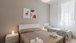 Un dormitorio con una cama grande, una cama más pequeña, una mesita de noche con una lámpara y una ventana con cortinas de gasa.