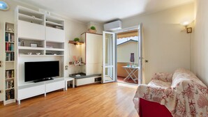 A living room with a flat-screen TV, white shelving, a red sofa, and a view through an open door to a balcony. - Apartment 'Raggio Di Sole' with Balcony and Wi-Fi (Torre del Lago Puccini)