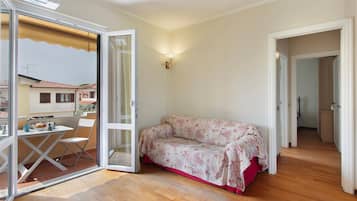 Una habitación con piso de madera, un sofá con funda floral, una mesa con tetera y tazas, y un vidrio corredizo que da a un área exterior.