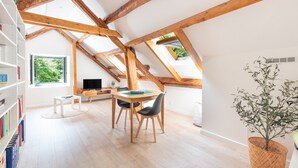 A room with a sloped ceiling, wooden beams, a dining area with a table and chairs, a bookshelf, and a window with a view of greenery. - Holiday Home 'L'authentik' with Mountain View, Private Terrace and Air Conditioning (Accous)