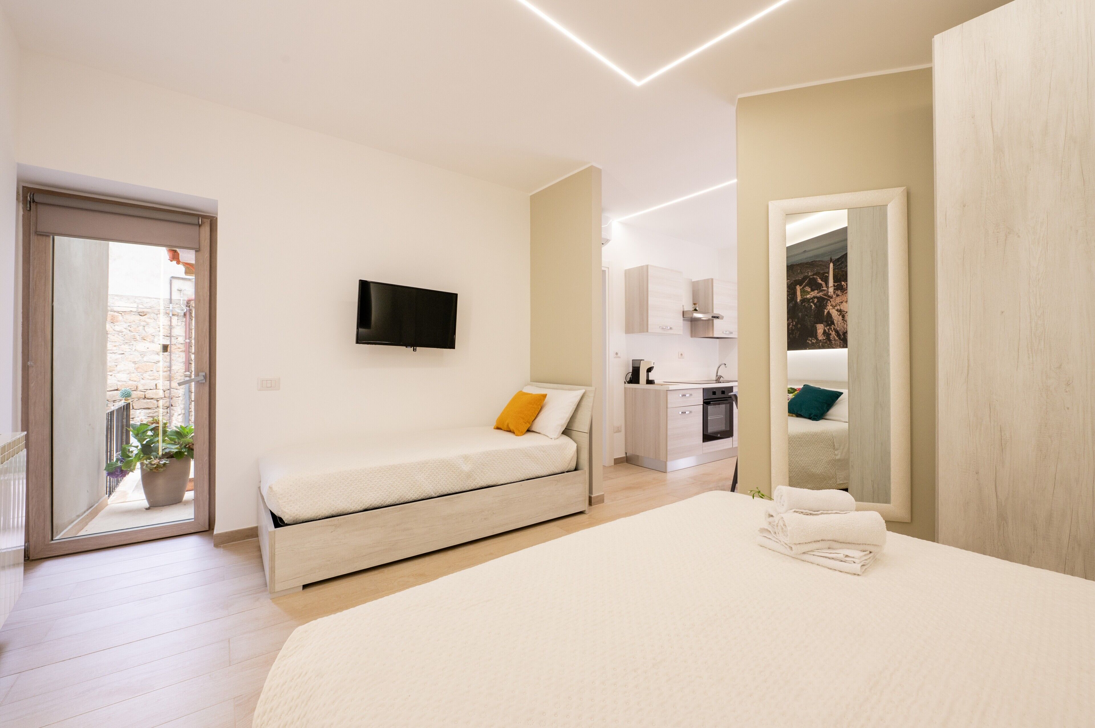 A modern bedroom with a bed, a TV, a small kitchen area, and a balcony.