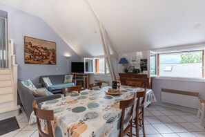 Uma sala de jantar com mesa posta para quatro pessoas, um sofá, uma televisão e uma janela com vista para o verde.