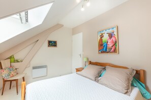 Quarto com teto inclinado, cama, cadeira, mesinha e um quadro na parede.