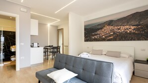 A modern hotel room with a grey sofa, a bed with white bedding, a dining area, and a cityscape view.
