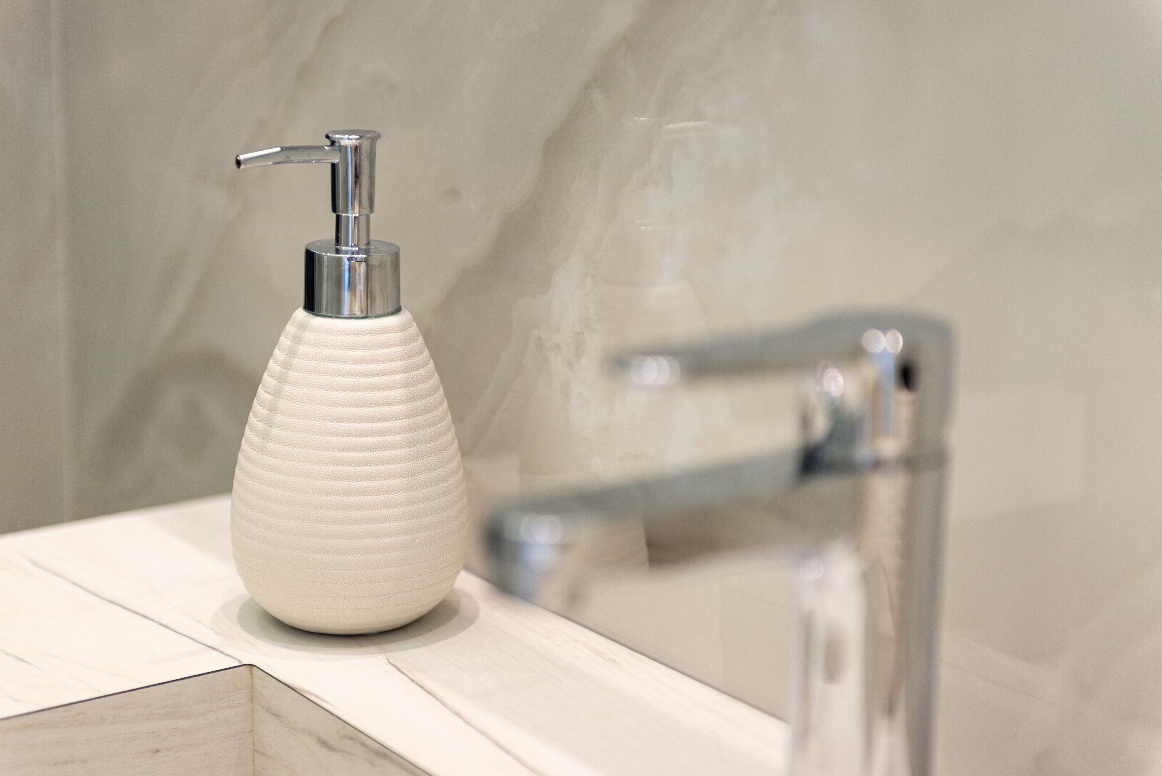 A bathroom with a white textured soap dispenser on a wooden surface, a chrome faucet, and a marble wall.
