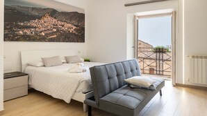 A bedroom with a bed, a grey sofa, a nightstand, a large window, and a cityscape painting.