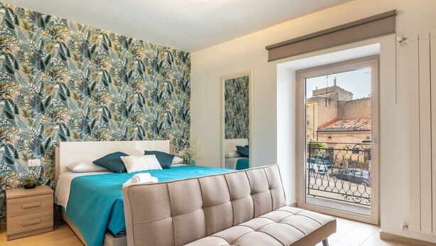 A bedroom with a bed, a sofa, a nightstand, a window with a view of buildings, and a patterned wall.