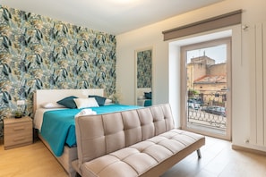 Une chambre à coucher comprenant un lit, un canapé, une table de chevet, une fenêtre donnant sur des immeubles et un mur décoré d’un motif.