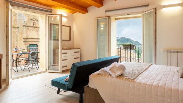 A bedroom with a bed, a desk, and a chair, a balcony with a view, and a sliding glass door leading to an outdoor area.