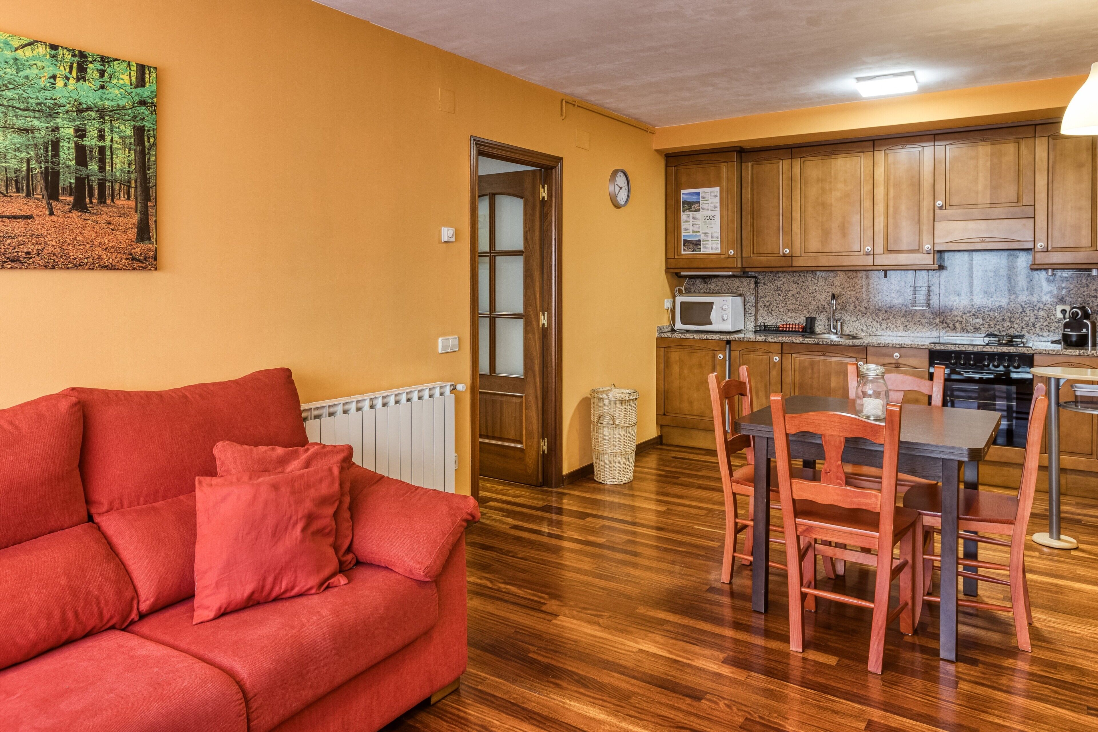 Ein Wohnzimmer mit einem roten Sofa, einem Esstisch mit Stühlen, Holzschränken, einer Mikrowelle und einer Wanduhr.