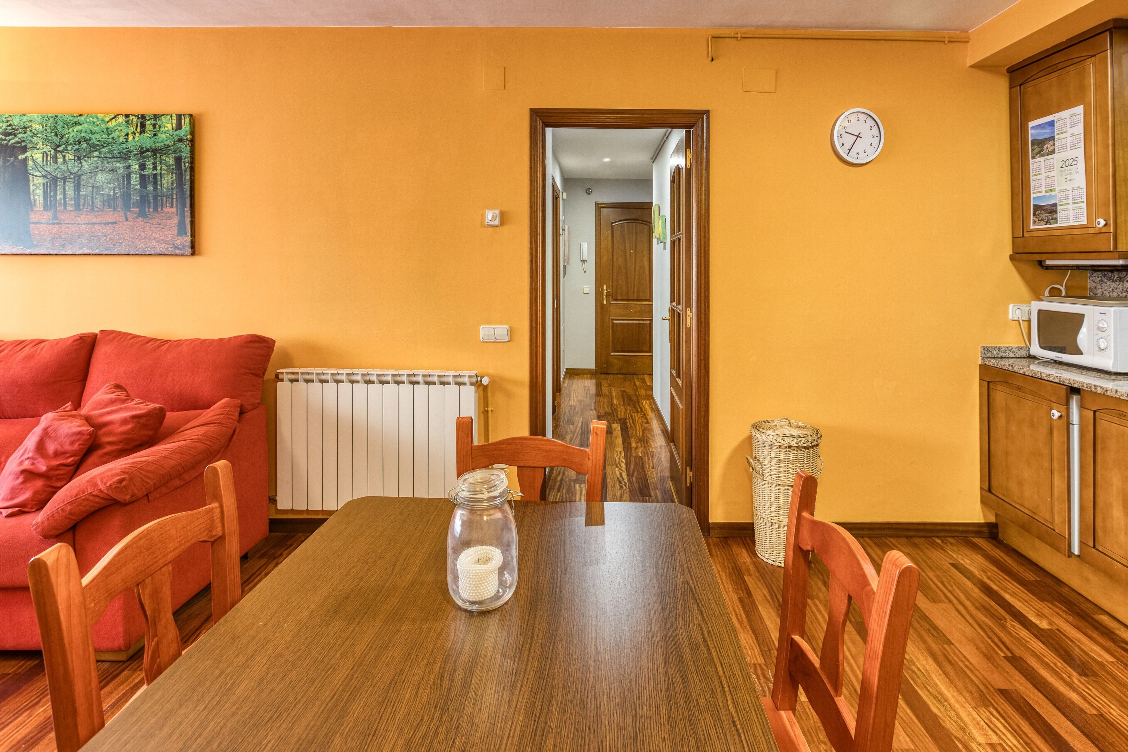 Un comedor con una mesa de madera, sillas, un sofá rojo, un cuadro, un reloj y una zona de cocina con microondas.
