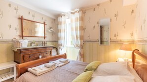 Ein Schlafzimmer mit Bett, Schminktisch, Spiegel, einem Fenster mit Vorhängen und einem Nachttisch mit Lampe.