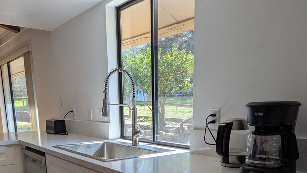 A modern kitchen with white cabinetry, a stainless steel sink, a coffee maker, and a window overlooking a garden. - Peaceful Three Rivers Retreat Near Sequoia National Park with EV charger, AC (Three Rivers)