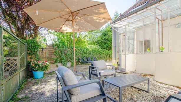 Een terras buiten met een tafel, stoelen, een grote parasol en een glazen constructie.