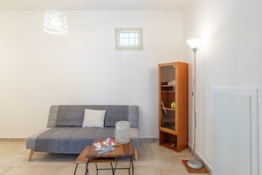 Un salon avec un canapé gris, une table d’appoint en bois, une lampe sur pied, une petite fenêtre et une bibliothèque.