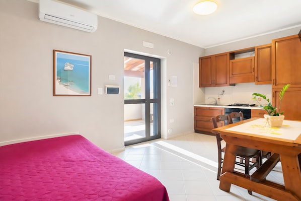A bedroom with a pink bedspread, a kitchen with wooden cabinets, and a dining area with a table and chairs.