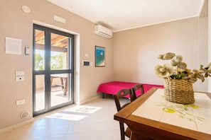 A room with a dining table, chairs, a bright pink bed, a woven basket with flowers, and a view through a glass door.