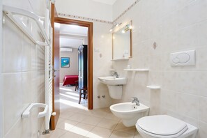 A modern bathroom with a toilet, bidet, sink, and mirror. There is a shower area visible in the background.