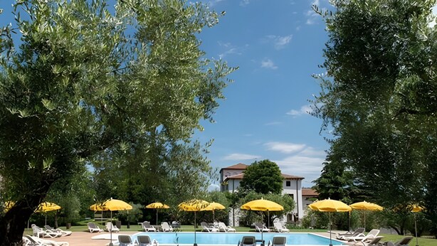 A poolside area with sun loungers and yellow umbrellas, surrounded by trees and a building in the background.