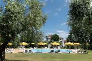 A poolside area with sun loungers and yellow umbrellas, surrounded by trees and a building in the background.