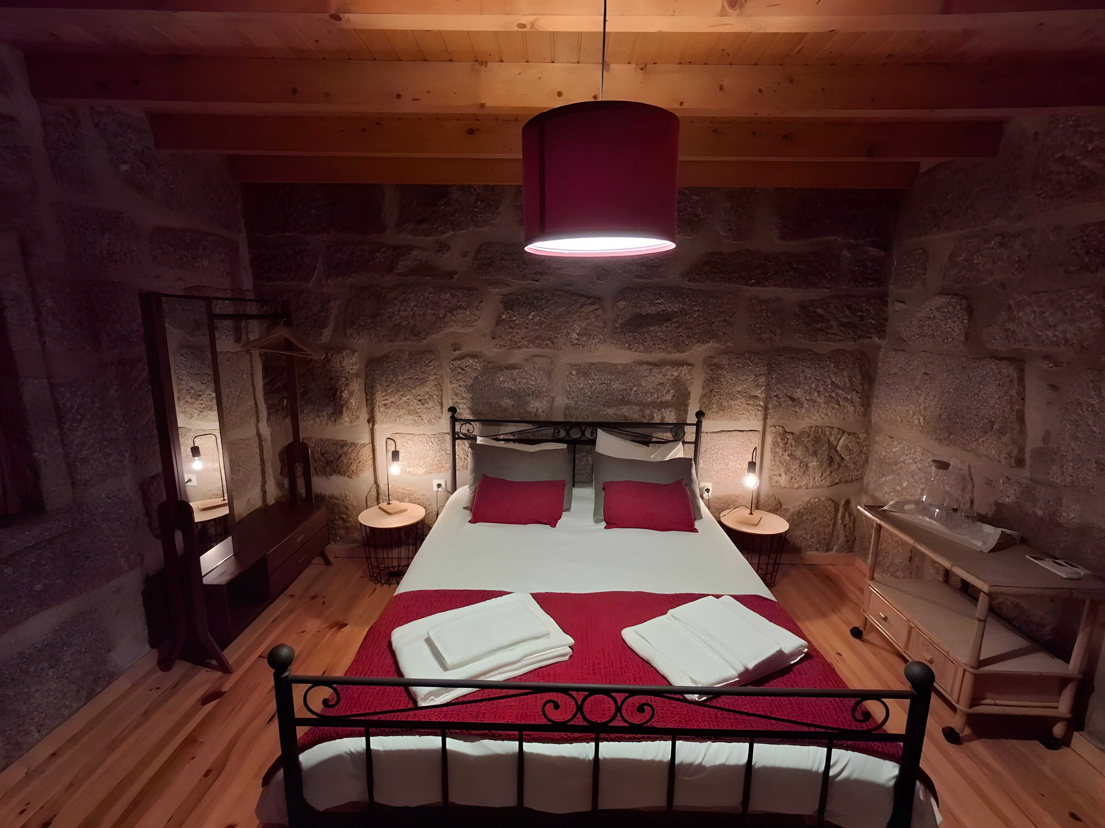 Une pièce aux murs de pierre et au plafond en bois, avec un lit recouvert d’une literie rouge, deux tables de chevet dotées de lampes et un luminaire suspendu.