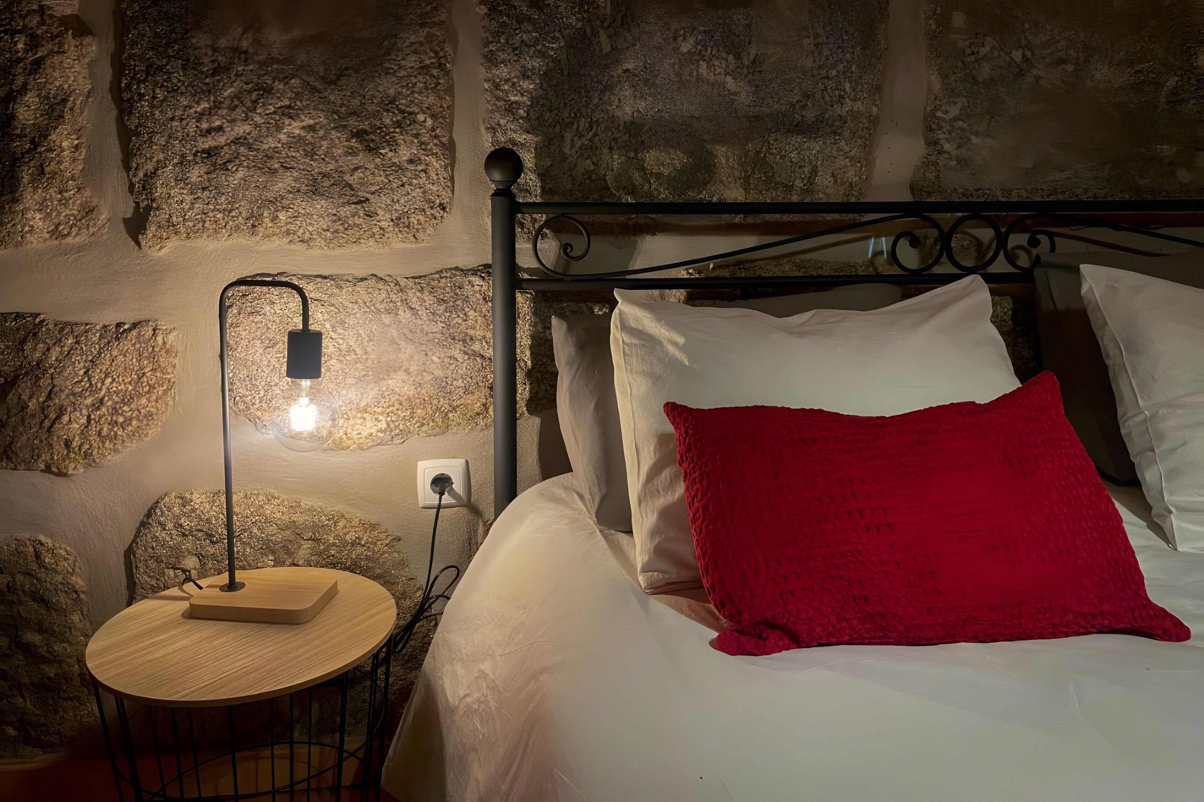 A room with a stone wall, a round wooden table with a lamp, and a bed with white bedding and a red pillow.