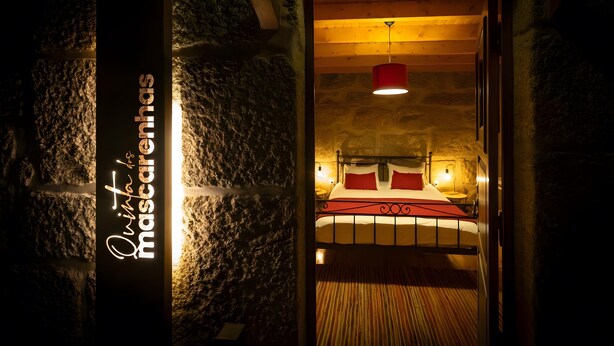 Habitación con paredes de piedra, una cama y una lámpara colgante roja, con un letrero que dice 'Quinta das Mascarinhas'.