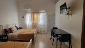 Ein Zimmer mit zwei Betten, einem Schreibtisch und Stühlen. An der Wand ist ein Fernseher angebracht, es gibt eine Klimaanlage und eine Tür mit Vorhang.