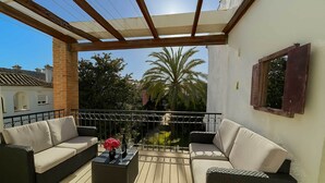 Zona salotto esterna coperta con due divani e un tavolino, una finestra con stipite in legno, e sullo sfondo palme e edifici.