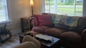 A living room with a brown leather sofa, a wooden coffee table, a lamp, and a view of a green landscape through large windows. - Charming 1-bedroom cottage in Holly Pond with AC, WiFi (Holly Pond)