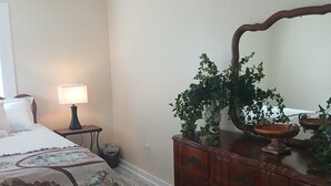 A bedroom with a bed, a nightstand with a lamp, a dresser with a mirror, and a potted plant. - Charming 1-bedroom cottage in Holly Pond with AC, WiFi (Holly Pond)