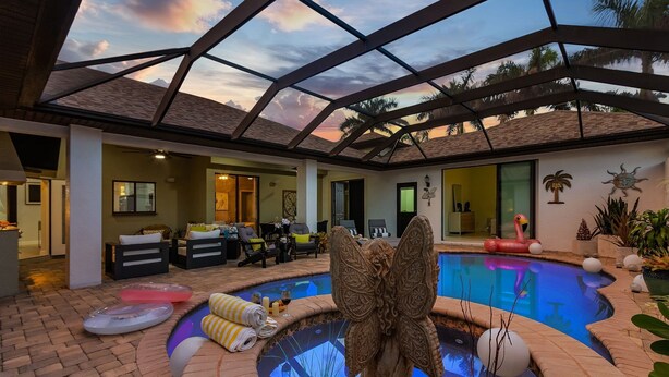 An indoor swimming pool with a butterfly sculpture, lounge chairs, and a sunset view through a glass ceiling. - Privacy, Luxury and Relaxation, Heated Pool & Spa - Villa Courtyard - Roelens (Cape Coral)