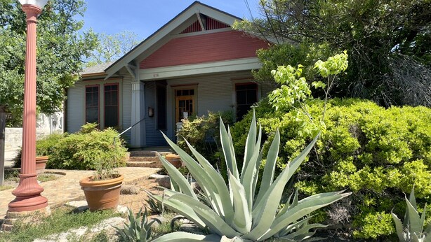 Une maison à deux étages, avec un toit rouge et un revêtement extérieur blanc, entourée d’un jardin où poussent de grands agaves et diverses espèces d’arbustes.