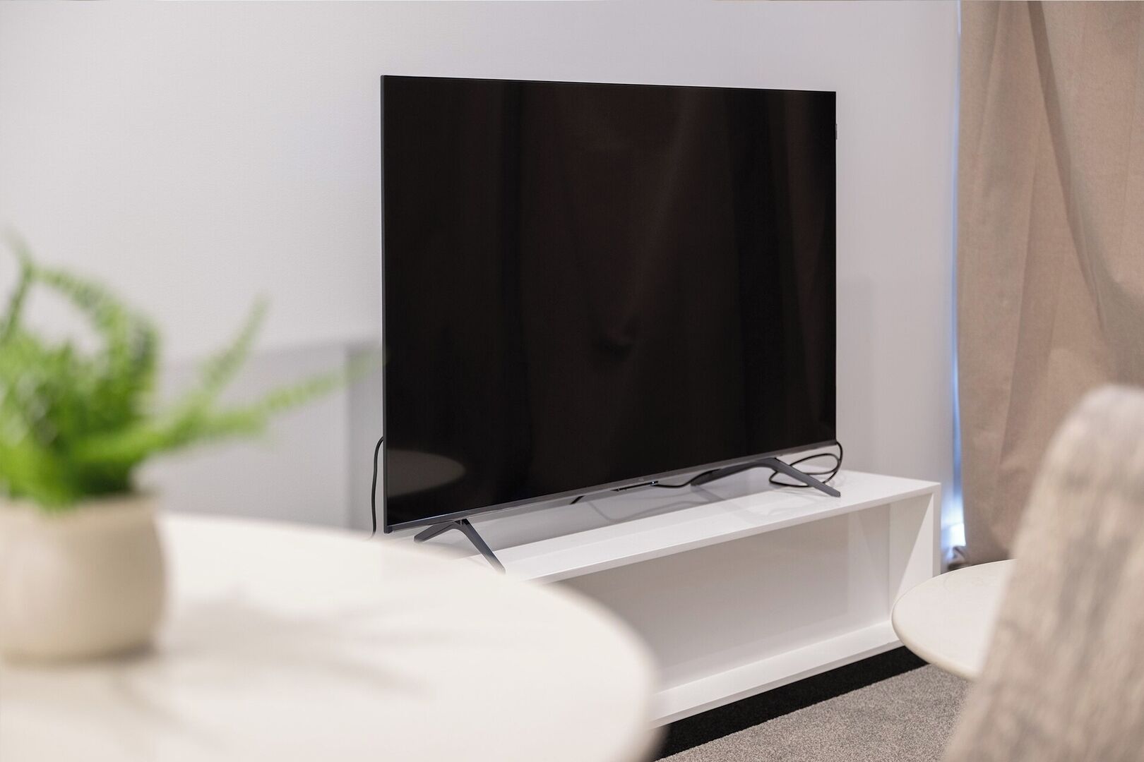 Un salon moderne avec un téléviseur à écran plat posé sur un meuble blanc, une petite table blanche et une plante en pot.