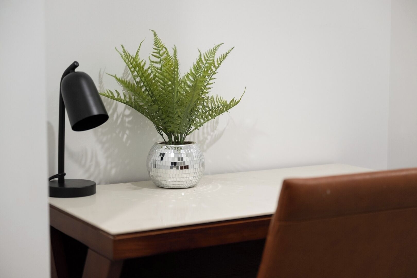 Uma escrivaninha com uma samambaia em vaso, uma luminária de mesa preta e uma cadeira de madeira.