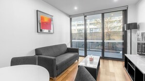 A modern living room with a grey sofa, a small round table, a flat-screen TV, and a large window with a view of a balcony and buildings.