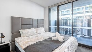 A modern bedroom with a large bed, a headboard, a bedside table with a lamp, and a view of a building through a balcony.