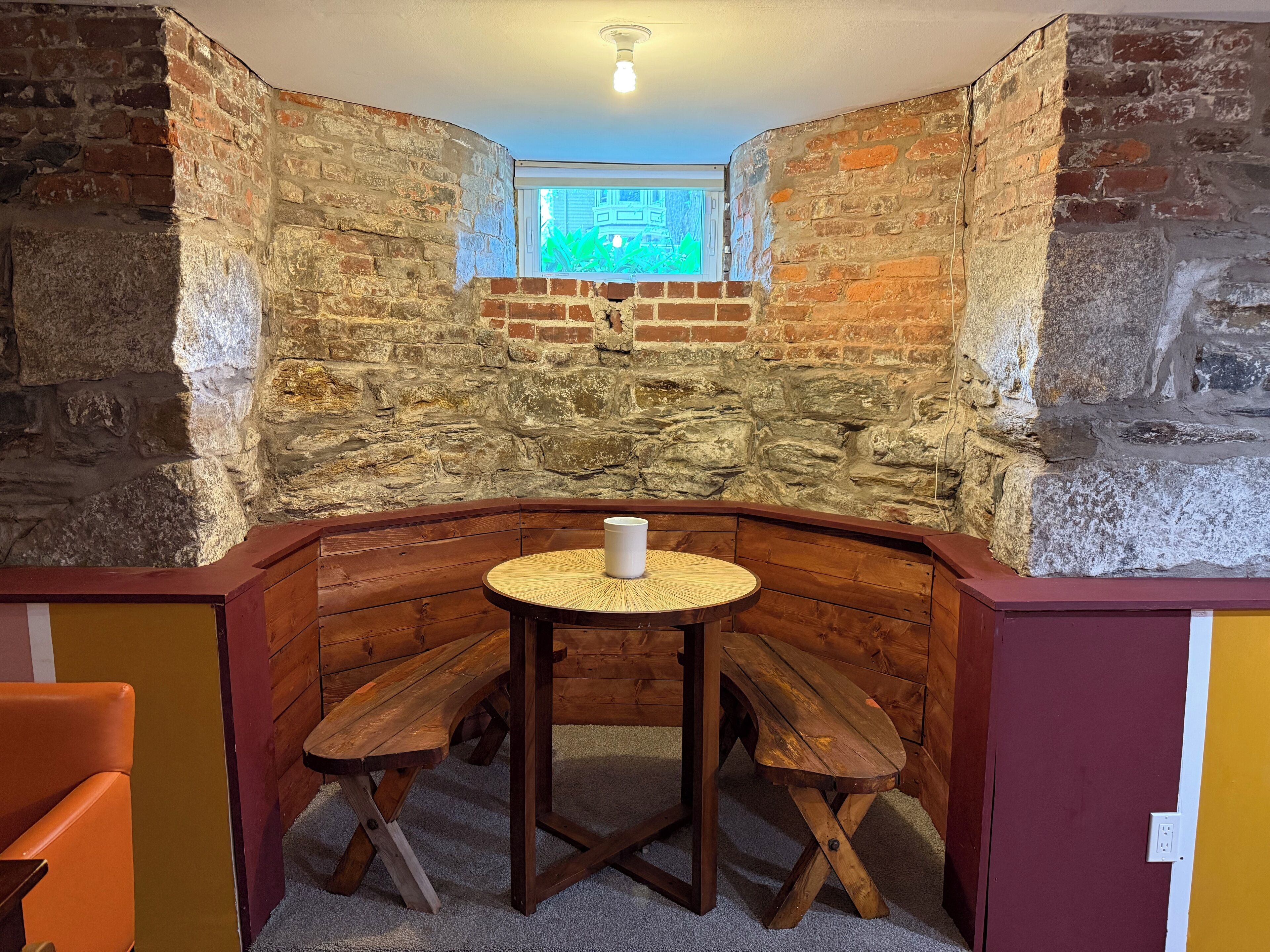 Un rincón amoblado con una mesa redonda de madera y bancos, una pared de piedra y una ventana con vistas.