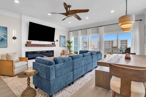 A modern living room with a sectional sofa, a flat-screen TV, a ceiling fan, and a dining area with a wooden table and chairs.