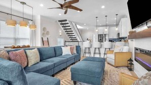 A modern living room with a blue sofa, ottoman, ceiling fan, and a staircase leading to the upper floor. - Ocean views-Steps from Beach-Elevator-3 Kings-Pets (Carolina Beach)