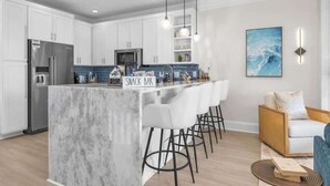 A modern kitchen with white cabinets, a marble island, bar stools, a refrigerator, and a microwave. - Ocean views-Steps from Beach-Elevator-3 Kings-Pets (Carolina Beach)
