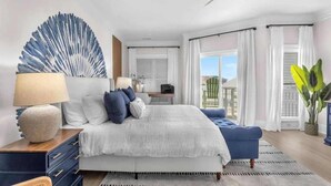 A bedroom with a bed, bedside tables, a lamp, a blue sofa, a large abstract wall art piece, and a view of the outdoors. - Ocean views-Steps from Beach-Elevator-3 Kings-Pets (Carolina Beach)