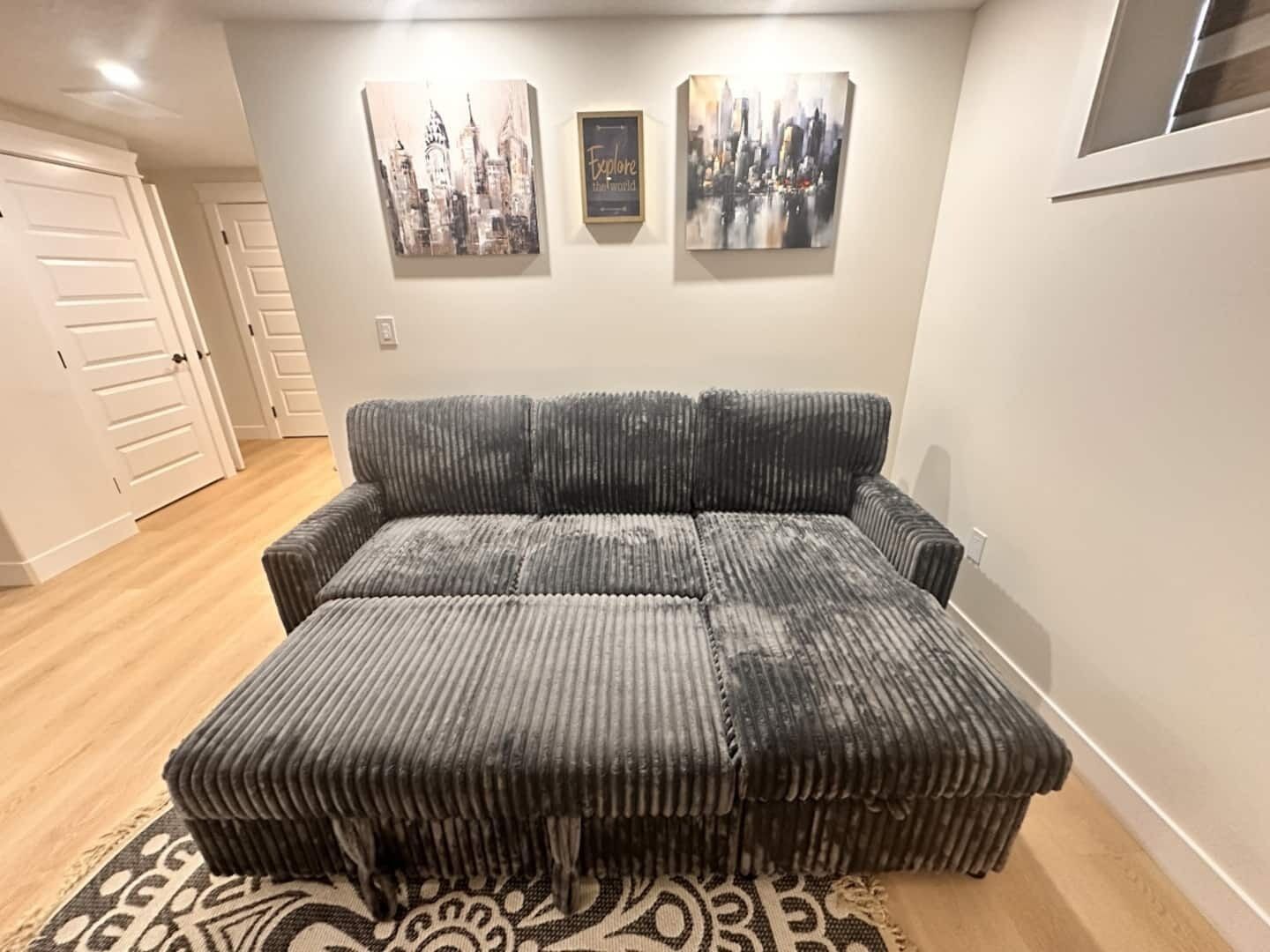 A living room with a grey sectional sofa and ottomans, a patterned rug, and two cityscape paintings on the wall.
