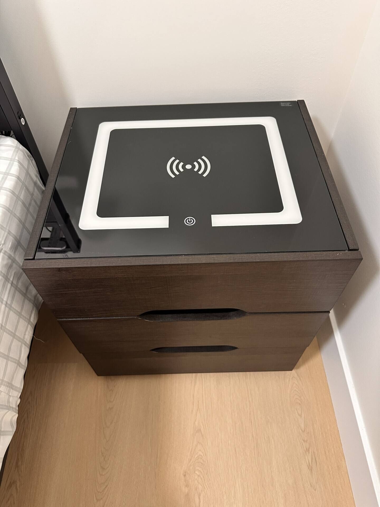 A compact, dark wooden bedside table with a flat-screen monitor, a power button, and a charging cable.