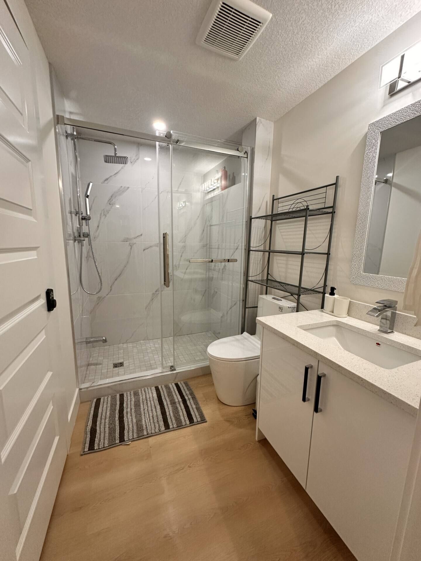A modern bathroom with a glass shower enclosure, a white toilet, a vanity with a mirror, and a wooden floor.