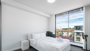 Una habitación de hotel moderna con una cama grande, una mesita de noche, un ventilador de pie y vistas a la ciudad a través de un ventanal amplio.