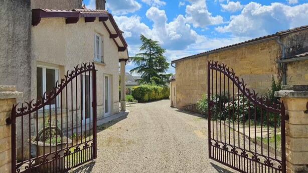 Une allée de gravier mène à une entrée fermée par un portail, avec une maison sur la gauche et un mur de pierre sur la droite.