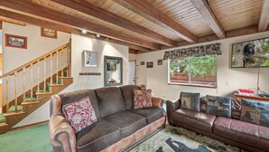 A spacious living room with a wooden staircase, leather sofas, and a large window with a view of trees.